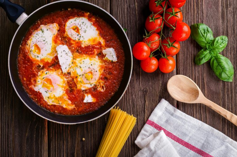 Delicious eggs in tomato sauce with basil, spaghetti, and fresh tomatoes on a wooden table.